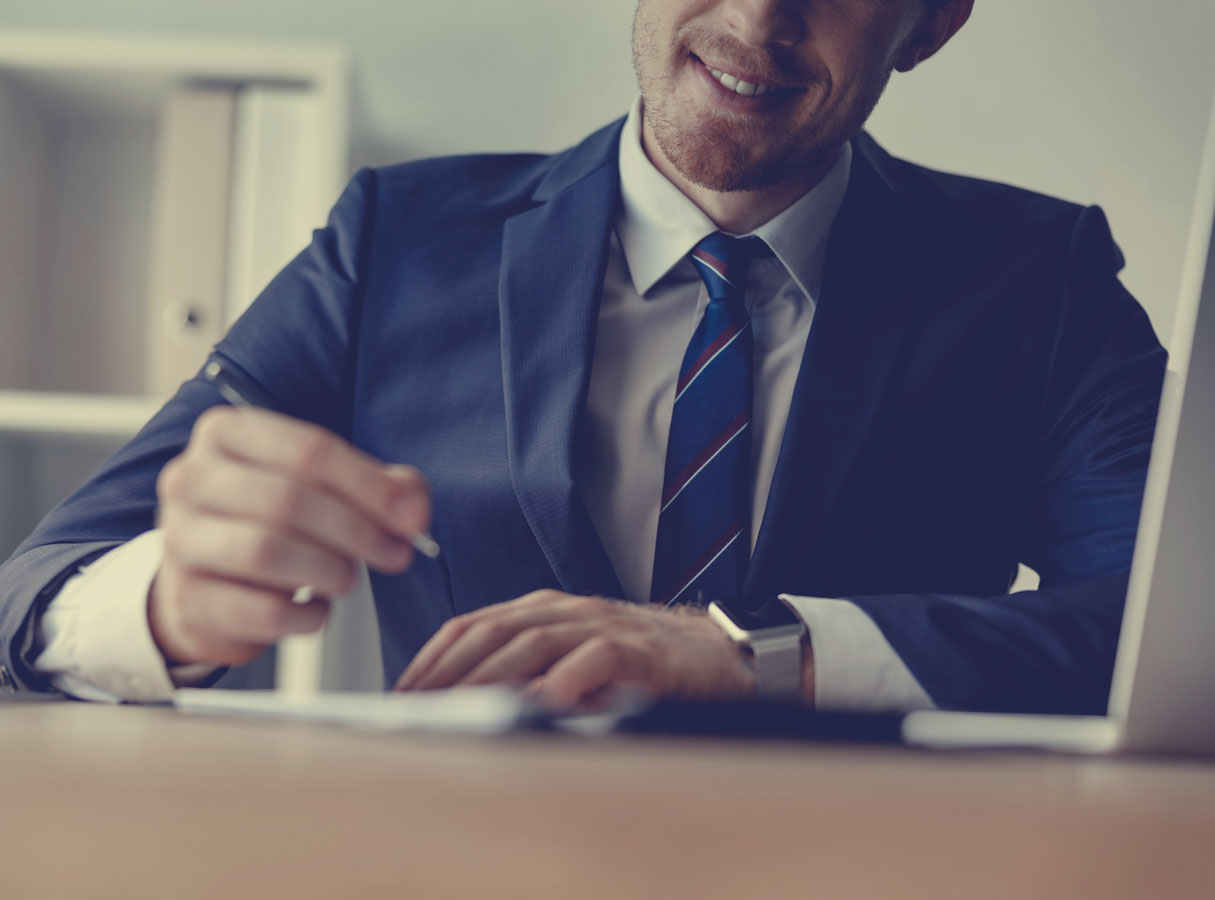 a smiling man in a suit and striped tie writes something down at a desk