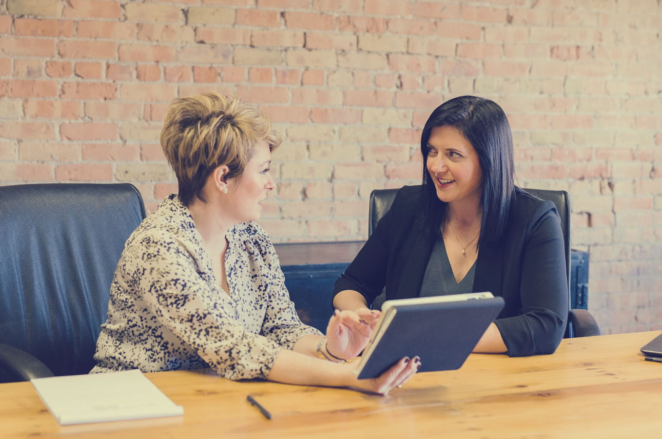 two women in business casual sitting at a table looking at a tablet