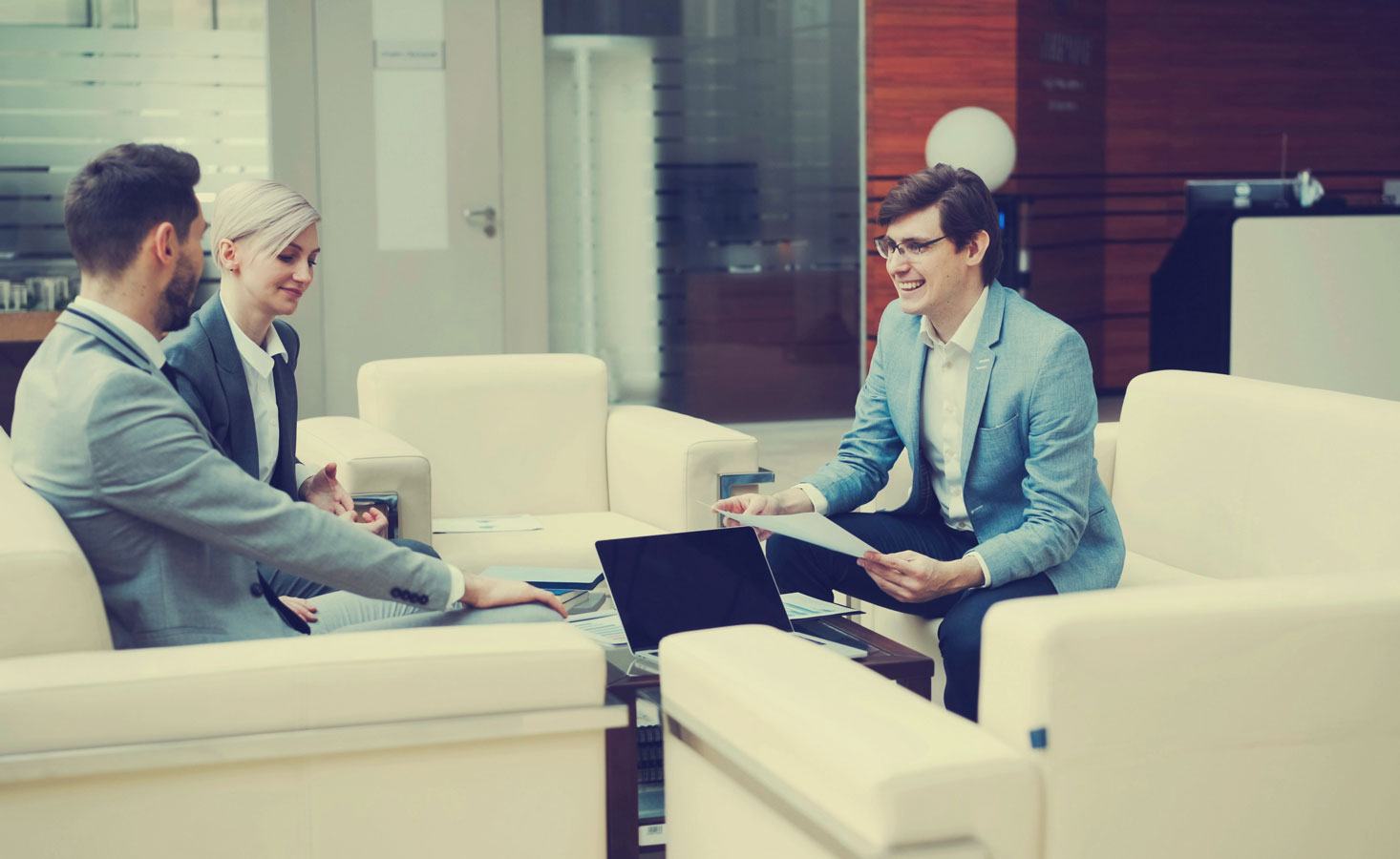 a group of people in suits sitting in armchairs with a laptop chatting and smiling